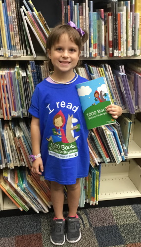 A young girl holding her program pamphlet and wearing a 1,000 books before kindergarten shirt